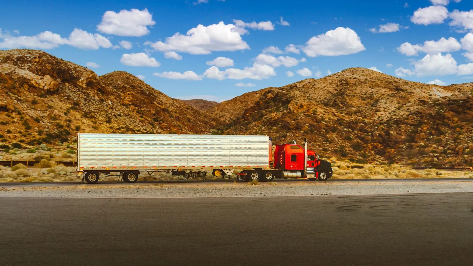 Red semi tractor pulling a white 53 foot dry van trailer along a desert highway with rocky hills behind it