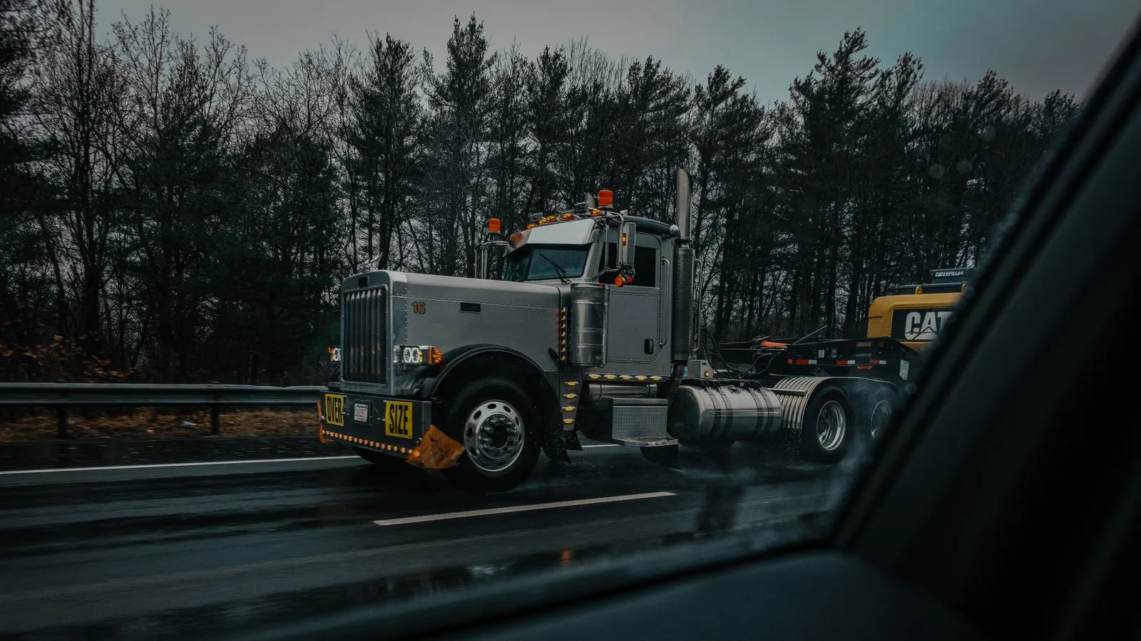 Gray semi tractor with an oversize load banner pulling a lowboy trailer carrying a yellow excavator on a wet highway